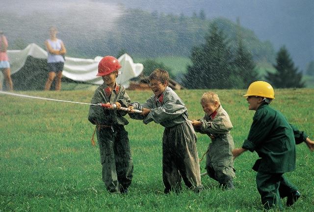 Vier Kinder mit Helmen und Overalls spritzen im Freien mit einem Schlauch Wasser, im Hintergrund sind zwei Erwachsene und eine Hügellandschaft zu sehen.