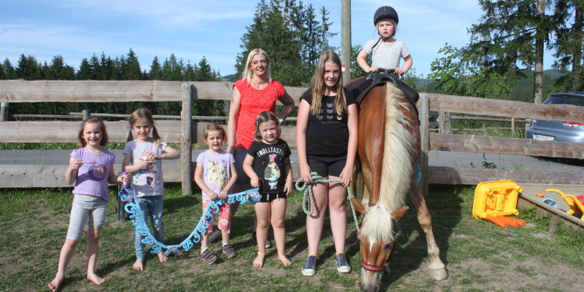 Sechs Kinder und ein Erwachsener posieren im Freien in der Nähe eines Holzzauns. Ein Kind sitzt auf einem Pony, während andere in der Nähe stehen und ein Seil und ein "Happy Birthday"-Schild halten. Im Hintergrund sind Bäume und Spielzeug zu sehen.