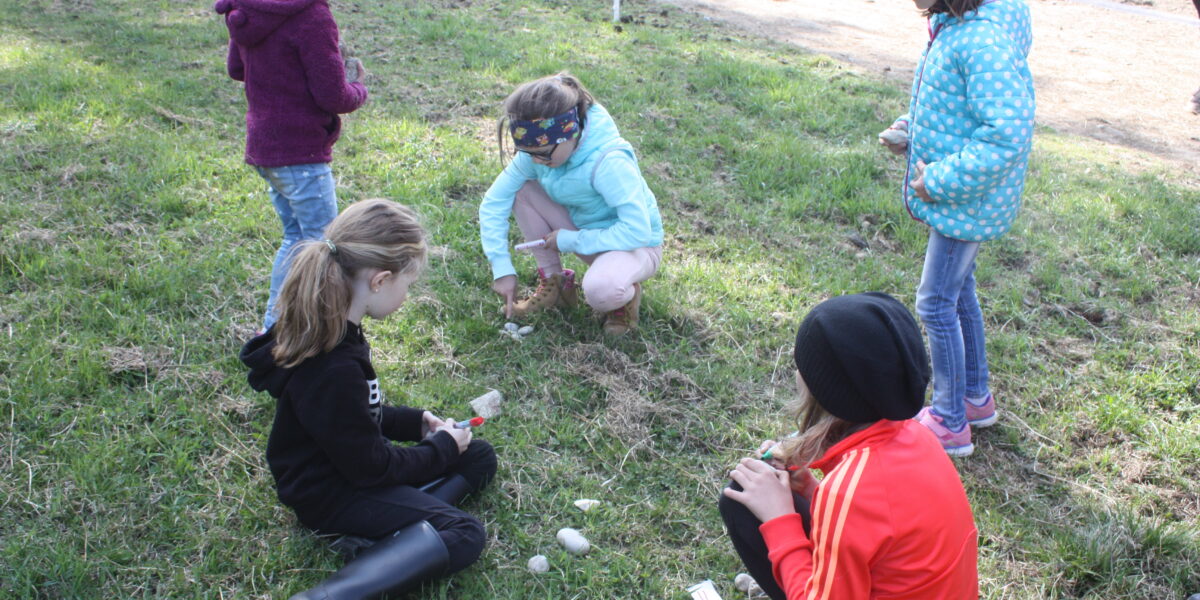 Fünf Kinder sitzen oder stehen im Freien auf einer Wiese und bemalen und verzieren Eier.