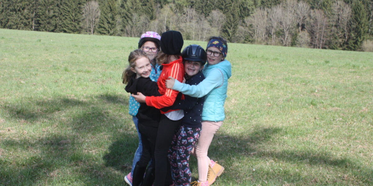 Sechs Kinder in legerer Outdoor-Kleidung und mit Helmen stehen in einer Gruppenumarmung auf einer Wiese, mit Bäumen und Hügeln im Hintergrund.
