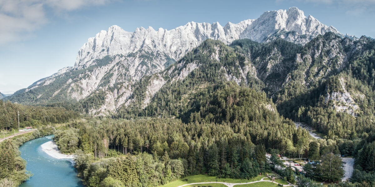 Ein gewundener Fluss fließt durch ein bewaldetes Tal mit einem kreisförmigen Garten vor der Kulisse hoher, felsiger Berge unter einem teilweise bewölkten Himmel.