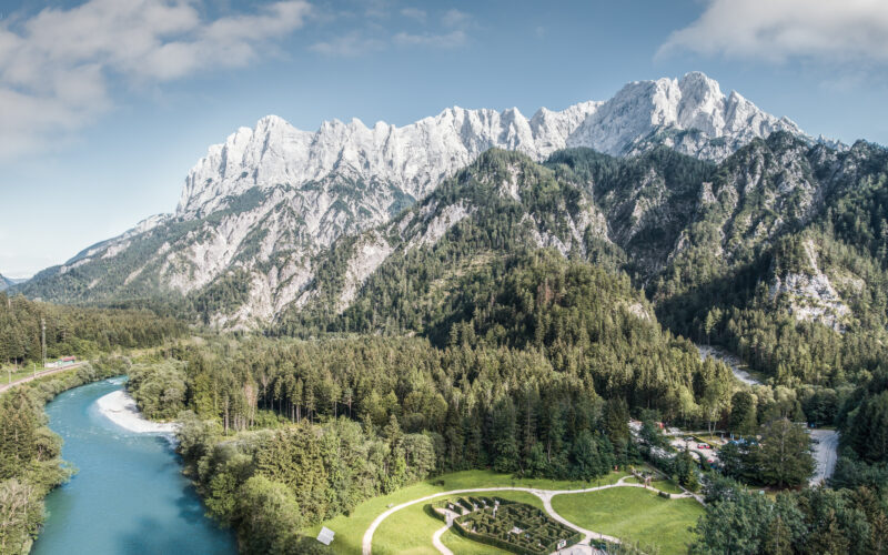 Ein gewundener Fluss fließt durch ein bewaldetes Tal mit einem kreisförmigen Garten vor der Kulisse hoher, felsiger Berge unter einem teilweise bewölkten Himmel.