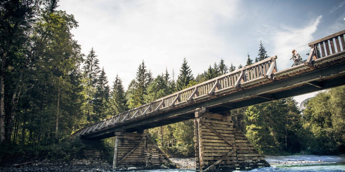 Eine Holzbrücke mit Geländer überspannt einen fließenden Fluss, umgeben von Bäumen und unter einem teilweise bewölkten Himmel.
