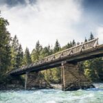 Eine Holzbrücke mit Geländer überspannt einen fließenden Fluss, umgeben von Bäumen und unter einem teilweise bewölkten Himmel.
