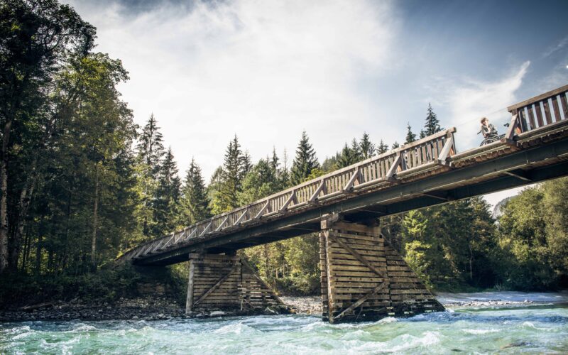 Eine Holzbrücke mit Geländer überspannt einen fließenden Fluss, umgeben von Bäumen und unter einem teilweise bewölkten Himmel.