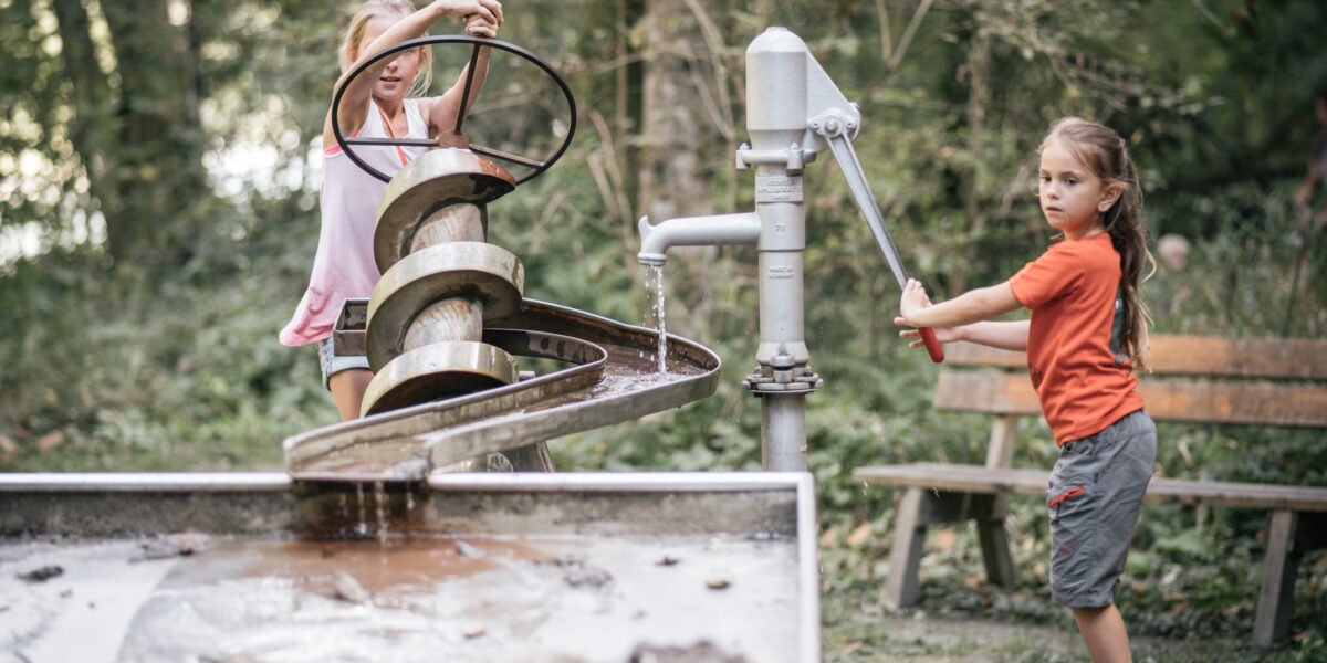 Zwei Kinder interagieren mit einer Wasserspielanlage im Freien. Das eine dreht eine Handpumpe, das andere bedient eine Archimedische Schraube. Im Hintergrund sind eine Holzbank und Bäume zu sehen.
