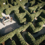 Luftaufnahme von vier Personen, die sich um eine Bank in der Mitte eines grünen Heckenlabyrinths versammelt haben, wobei Kieswege die Hecken trennen.