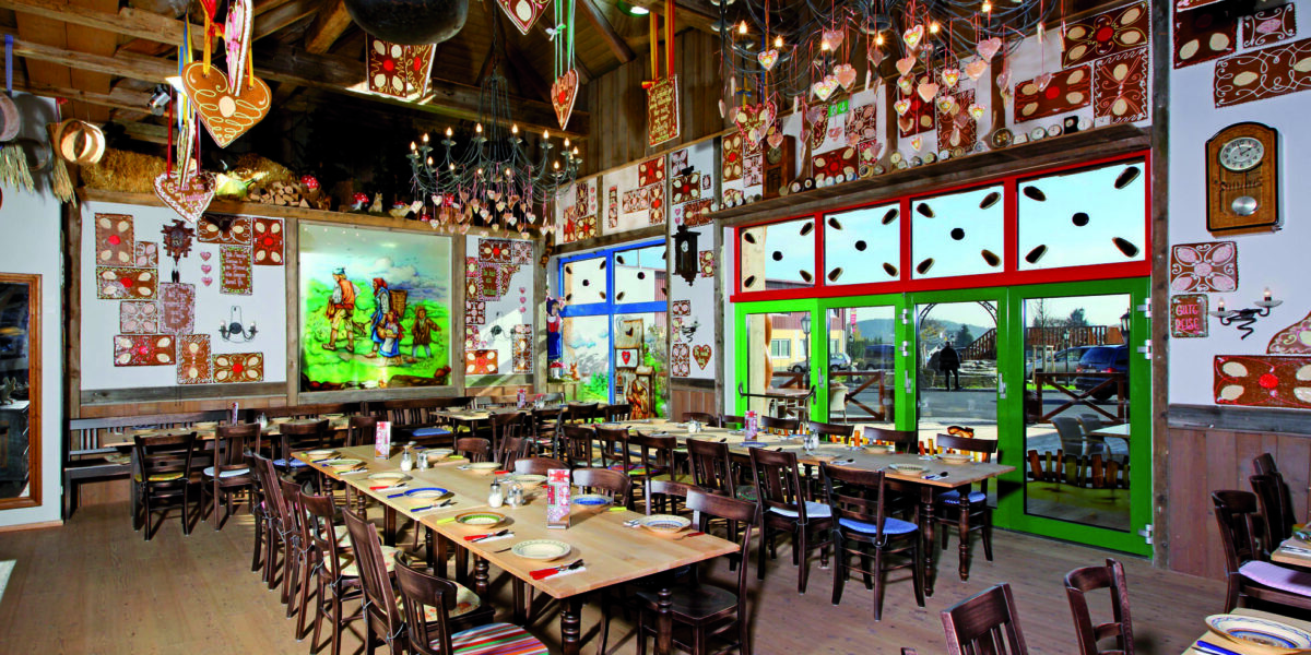 Das farbenfrohe Interieur des Restaurants mit langen Holztischen, uneinheitlichen Stühlen, farbenfroher Wandkunst, dekorativer Beleuchtung und großen, grün umrahmten Fenstern, die natürliches Licht hereinlassen.