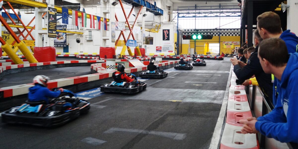 Indoor-Go-Kart-Rennfahrer fahren um eine Strecke, während die Zuschauer hinter einer Absperrung zuschauen; bunte Fahnen und Banner hängen über der Strecke.