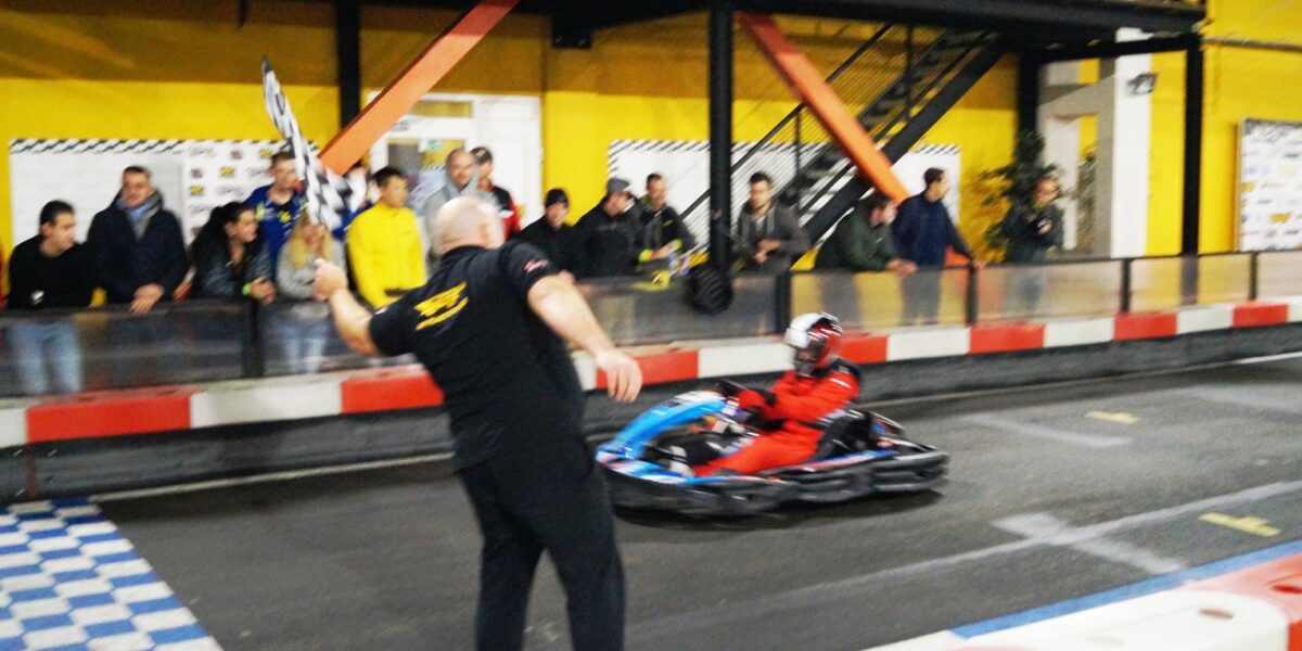 Eine Person winkt mit der Zielflagge, während ein Go-Kart-Fahrer in Rot die Ziellinie auf einer Indoor-Kartbahn überquert, während die Zuschauer hinter einer Absperrung zusehen.