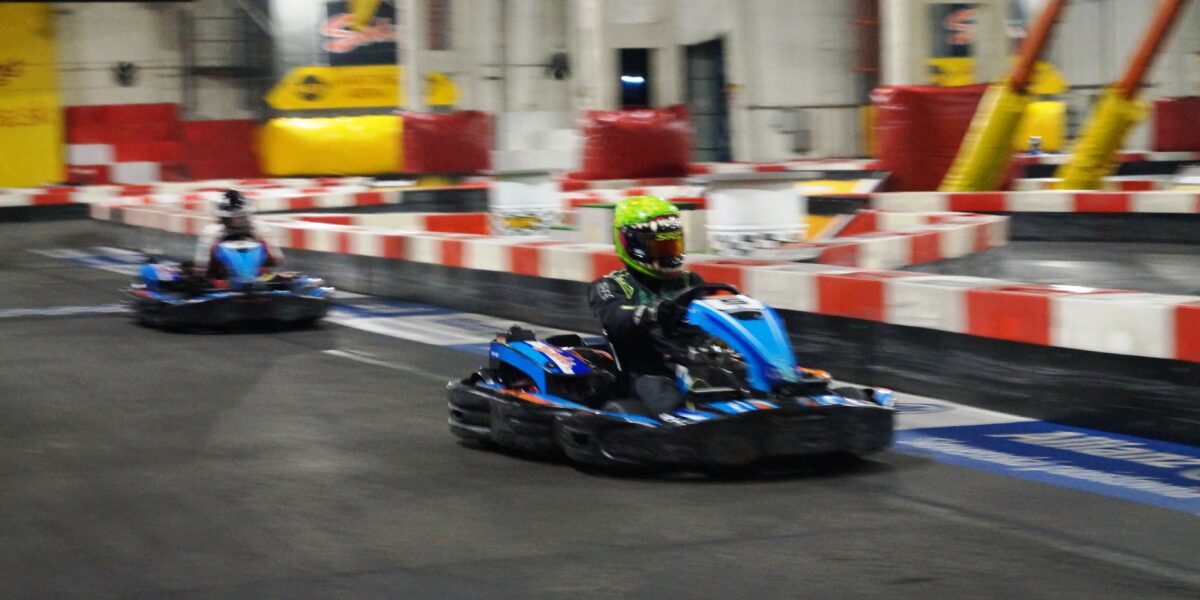 Zwei Personen fahren blaue Gokarts auf einer überdachten Rennstrecke mit roten und weißen Hindernissen.