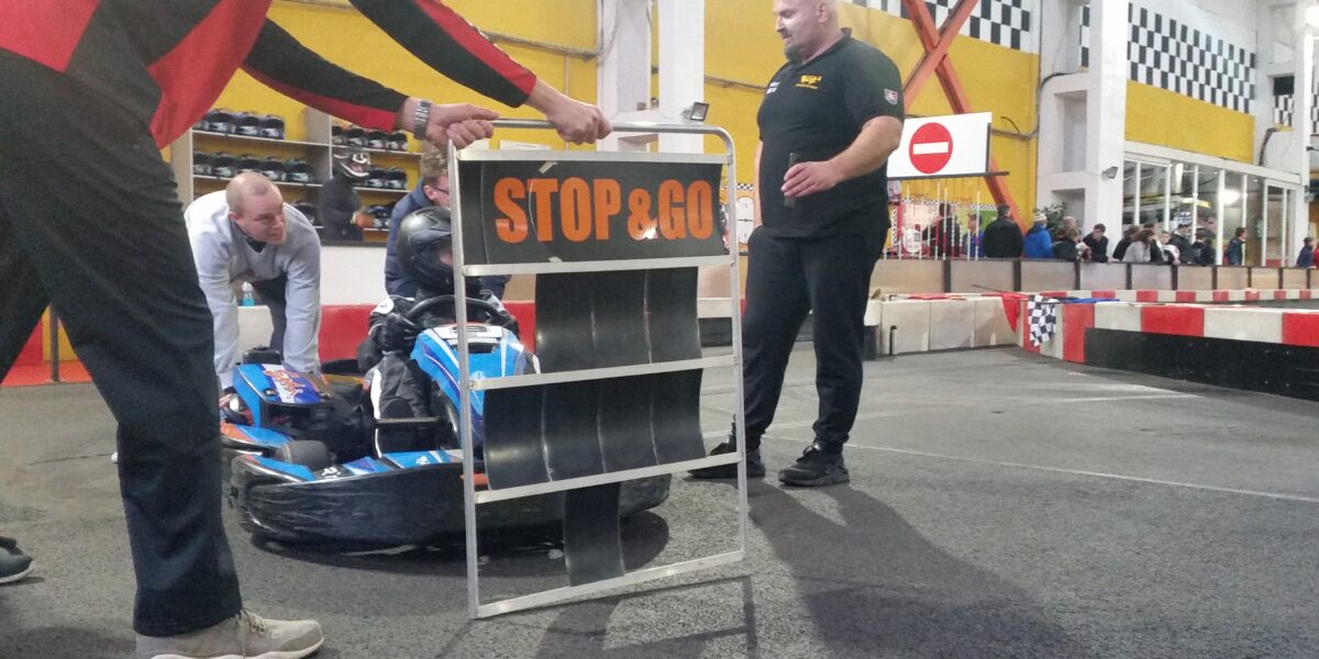 Eine Person in einem Go-Kart wird von einem Mitarbeiter mit einem "STOP & GO"-Schild auf einer Indoor-Bahn angehalten, während im Hintergrund weitere Personen und Absperrungen zu sehen sind.