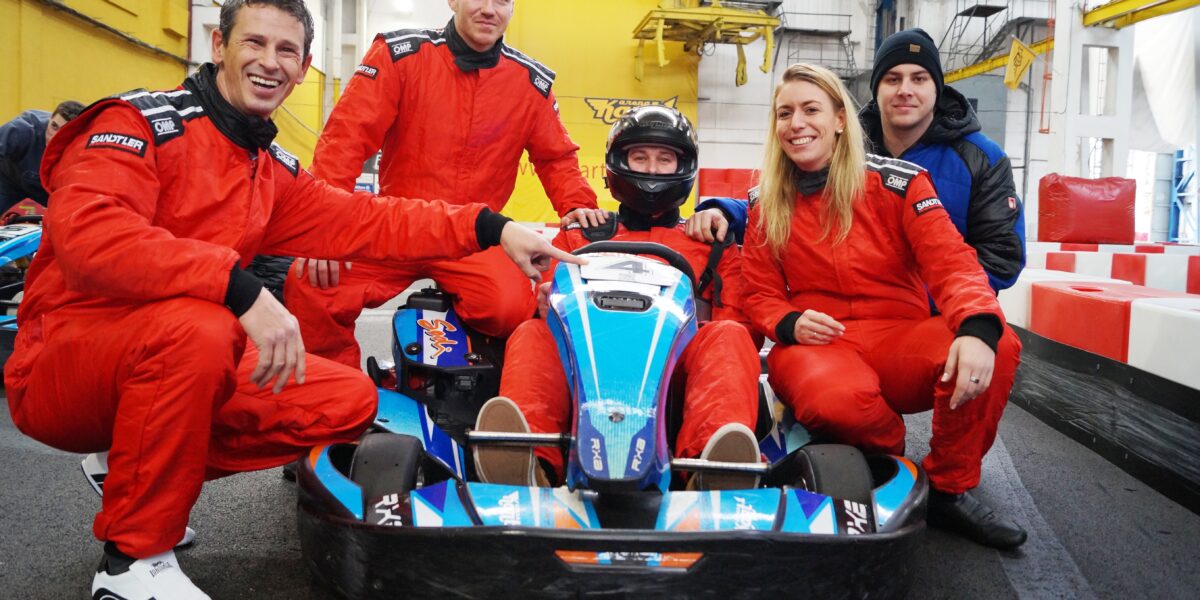 Fünf Personen in roten Rennanzügen posieren in einem Innenraum um ein blaues Go-Kart; eine Person sitzt im Kart und trägt einen Helm, während die anderen in der Nähe hocken oder sitzen und lächeln.