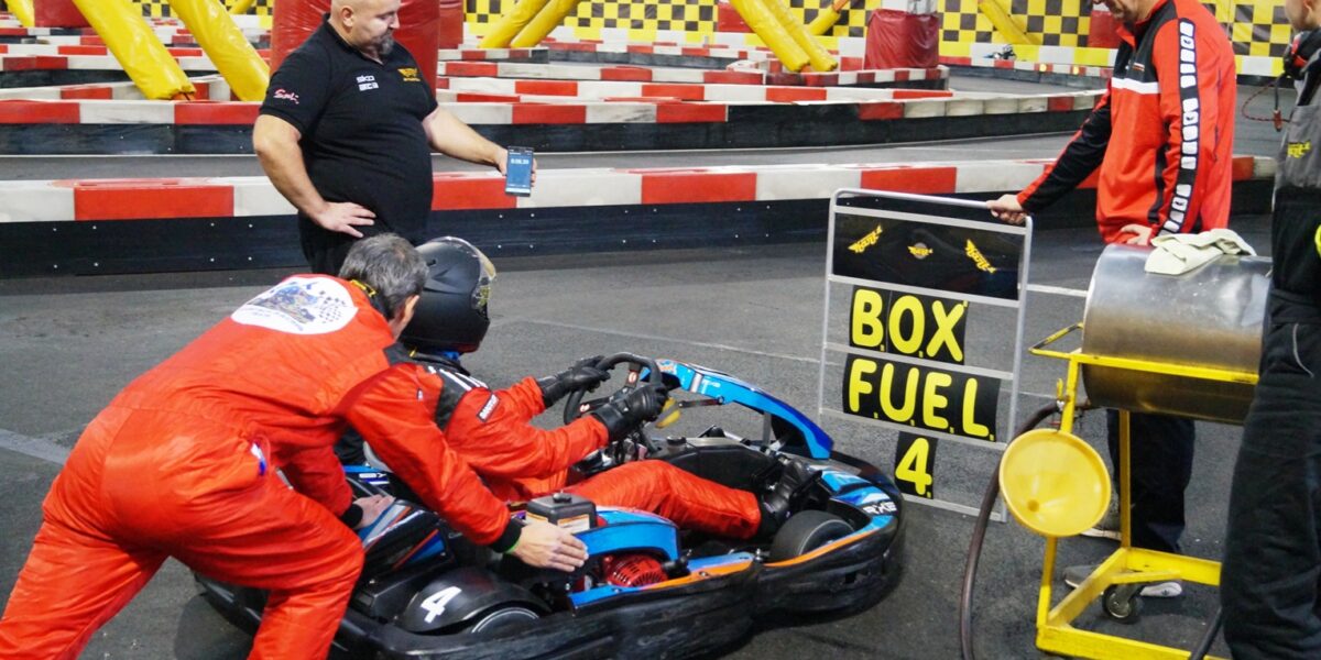 Zwei Personen schieben ein Go-Kart mit einem Fahrer in voller Montur, während drei Besatzungsmitglieder in der Nähe stehen; auf einem Schild steht "BOX FUEL 4" auf einer Indoor-Kartbahn.