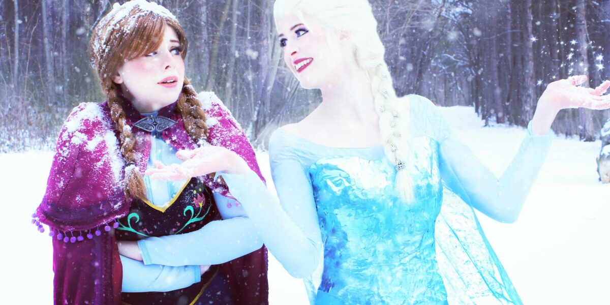 Zwei Frauen in Kostümen stehen in einem verschneiten Wald, wobei die eine ein blaues Kleid und die andere einen lila Umhang trägt. Schneeflocken fallen um sie herum, während sie sich unterhalten.