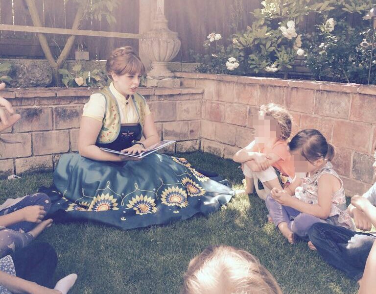 Eine Frau in einem grünen Kleid liest einer Gruppe von Kindern, die im Gras eines Gartens sitzen, aus einem Buch vor.