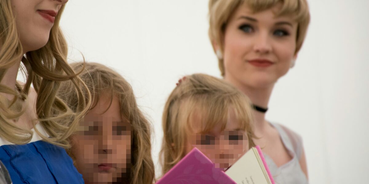 Zwei als Prinzessinnen verkleidete Erwachsene halten ein rosafarbenes Buch in der Hand und lesen zwei kleinen Kindern vor, deren Gesichter verpixelt sind.