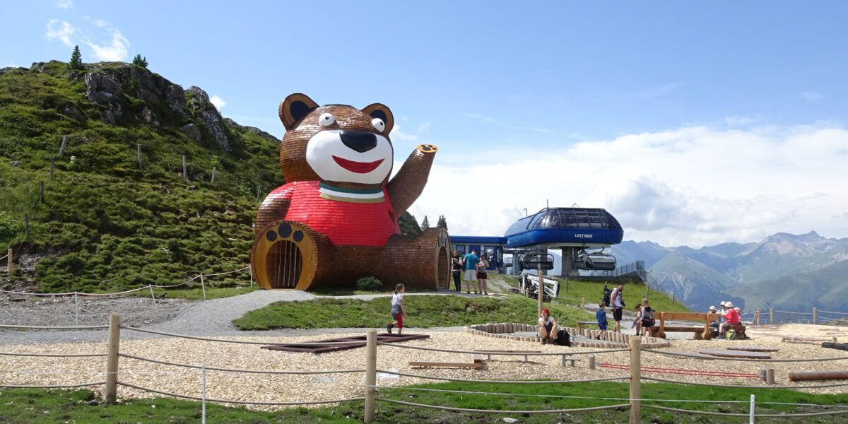 Ein großes bärenförmiges Gebäude und eine Skiliftstation befinden sich auf einem grasbewachsenen Berghang mit einem eingezäunten Schotterspielplatz und fernen Bergen im Hintergrund.