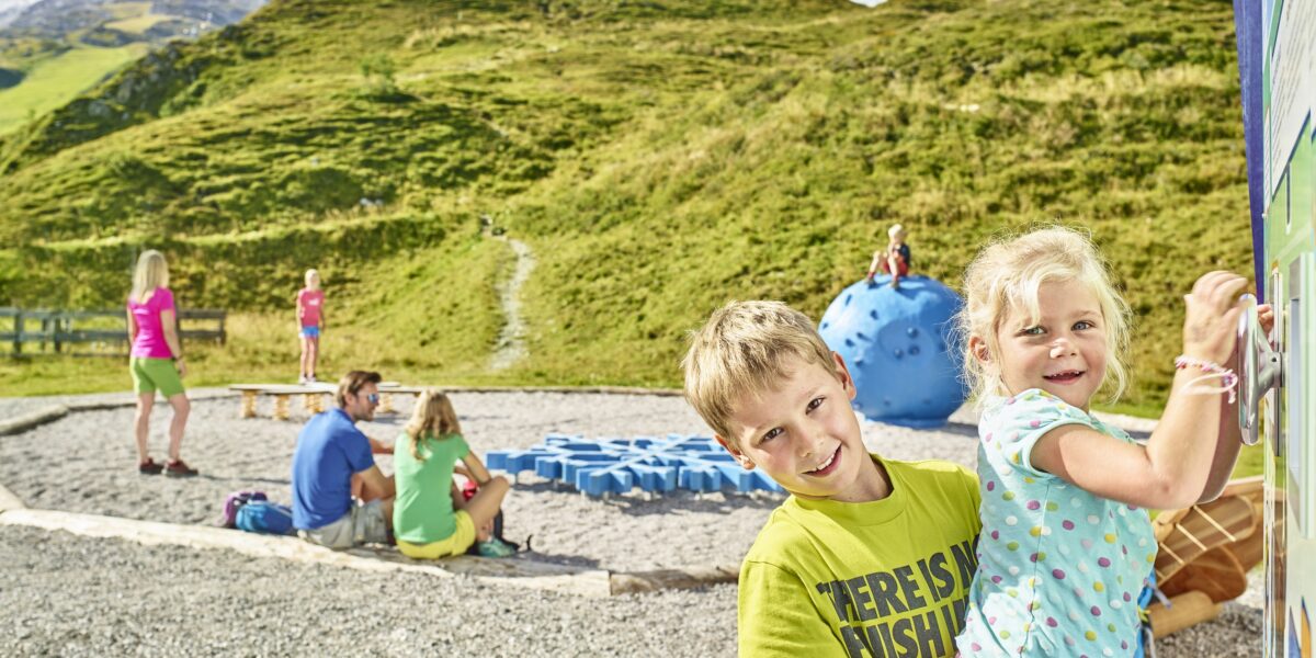 Kinder spielen auf einem Spielplatz im Freien in einer bergigen Gegend mit grünen Hügeln und felsigen Gipfeln im Hintergrund unter einem klaren Himmel.