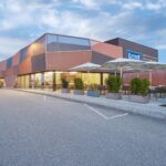 Modernes Geschäftsgebäude mit geometrisch gemusterter Fassade, großen Fenstern und einem Sitzbereich im Freien mit Sonnenschirmen und Pflanzgefäßen, angrenzend an eine Straße.