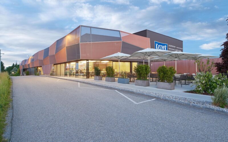 Modernes Geschäftsgebäude mit geometrisch gemusterter Fassade, großen Fenstern und einem Sitzbereich im Freien mit Sonnenschirmen und Pflanzgefäßen, angrenzend an eine Straße.