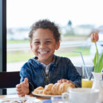 Lächelndes Kind mit Milchschnurrbart, das an einem Tisch mit Gebäck, Saft und einer Blumenvase in einem hellen Café sitzt.