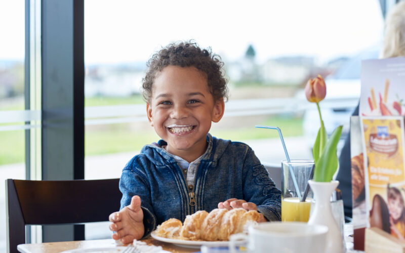 Lächelndes Kind mit Milchschnurrbart, das an einem Tisch mit Gebäck, Saft und einer Blumenvase in einem hellen Café sitzt.