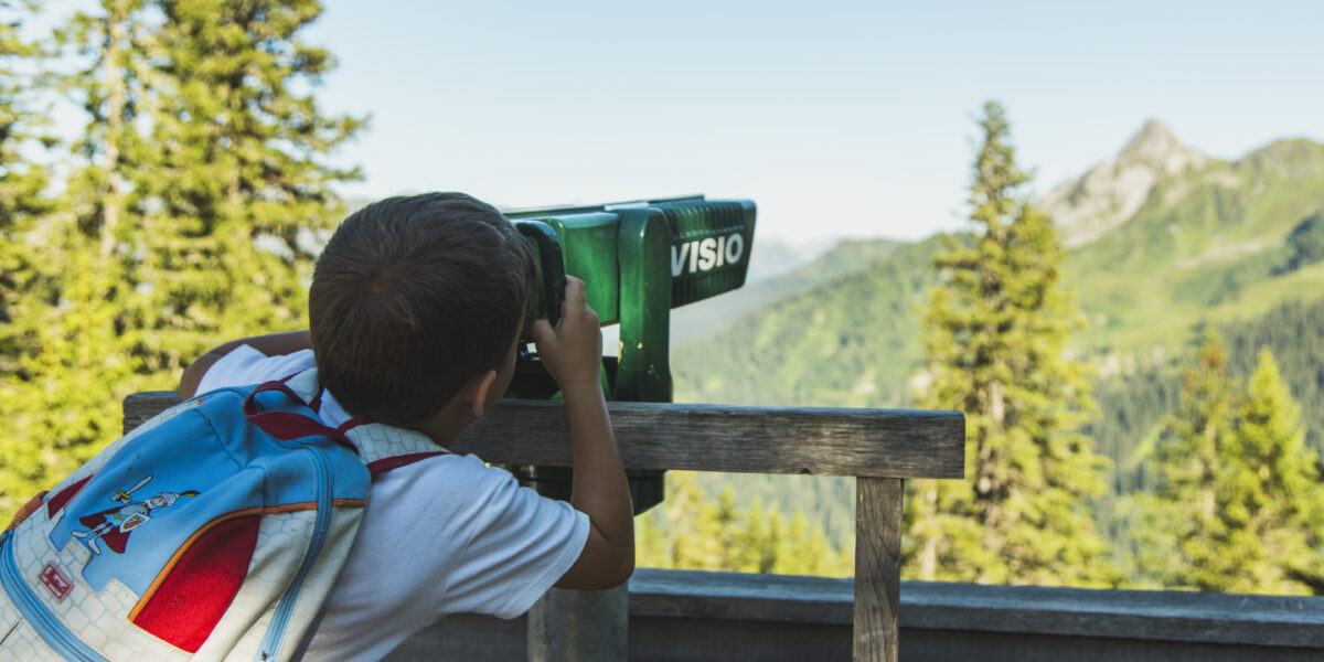 Ein Kind mit einem blauen Rucksack blickt durch einen grünen Landschaftsbetrachter auf eine von Bäumen umgebene Berglandschaft.