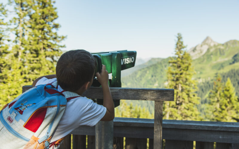 Ein Kind mit einem blauen Rucksack blickt durch einen grünen Landschaftsbetrachter auf eine von Bäumen umgebene Berglandschaft.