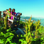 Eine Gruppe von Kindern steht auf einer kleinen Holzbrücke über einen Bach, umgeben von grünen Pflanzen und Bergen unter einem klaren blauen Himmel.