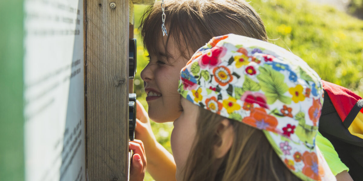Zwei Kinder schauen sich an einem sonnigen Tag eine Informationstafel im Freien genau an; das eine trägt einen bunten Blumenhut, das andere blinzelt zum Lesen.