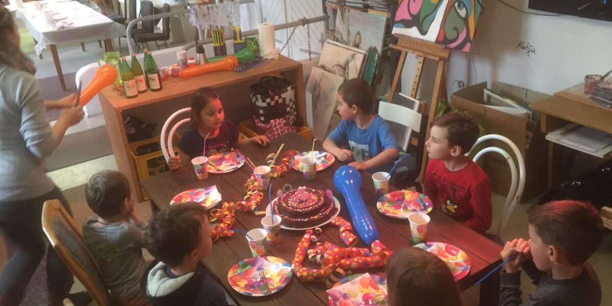 Eine Gruppe von Kindern sitzt um einen Tisch mit Kuchen, Tellern und Snacks in einem mit Gemälden dekorierten Kunstatelier. Ein Erwachsener steht in der Nähe und hält Luftballons.