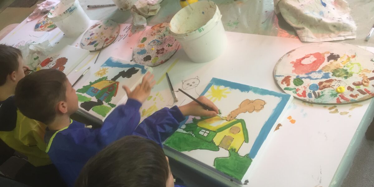 Die Kinder sitzen an einem Tisch und malen mit Pinsel und Palette bunte Bilder von Häusern und Naturszenen.