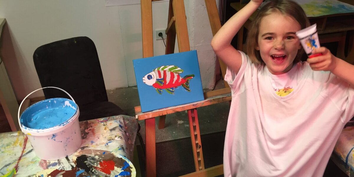 Ein lächelndes junges Mädchen in einem weißen Hemd hält Pinsel und Farbtube hoch und steht neben einem Fischbild auf einer Staffelei in einem Kunstatelier. Auf dem Tisch liegen Malutensilien und eine Farbpalette.