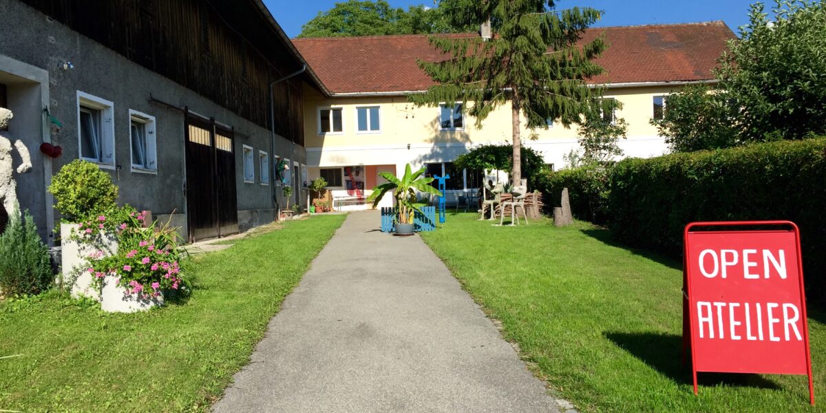 Ein gepflasterter Weg führt zu einem gelben Gebäude mit einem roten Schild auf dem Rasen, auf dem "OPEN ATELIER" steht; Topfpflanzen säumen den Weg.