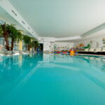 Hallenbad mit klarem, blauem Wasser, Palmen an einer Seite, Liegestühlen und farbenfrohen Wandmalereien mit Wasserthemen im Hintergrund.