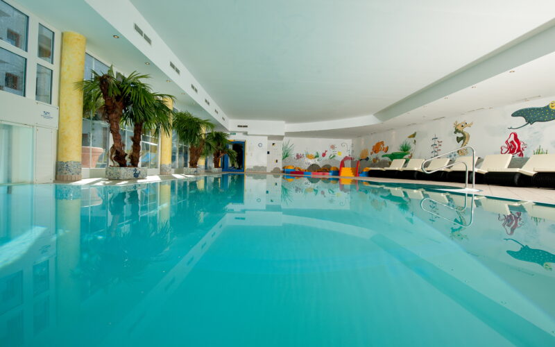 Hallenbad mit klarem, blauem Wasser, Palmen an einer Seite, Liegestühlen und farbenfrohen Wandmalereien mit Wasserthemen im Hintergrund.