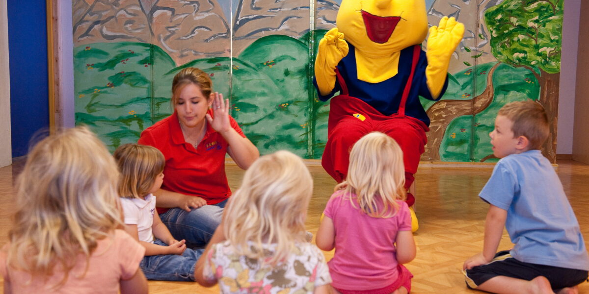 Eine Gruppe kleiner Kinder sitzt auf dem Boden um eine Frau und eine Person in einem gelben Maskottchenkostüm in einem Spielzimmer mit einer gemalten Bergkulisse.