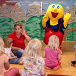 Eine Gruppe kleiner Kinder sitzt auf dem Boden um eine Frau und eine Person in einem gelben Maskottchenkostüm in einem Spielzimmer mit einer gemalten Bergkulisse.