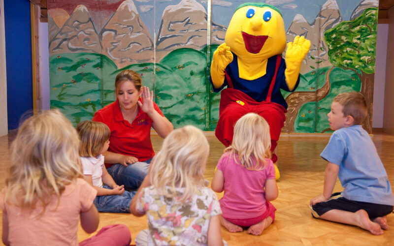 Eine Gruppe kleiner Kinder sitzt auf dem Boden um eine Frau und eine Person in einem gelben Maskottchenkostüm in einem Spielzimmer mit einer gemalten Bergkulisse.