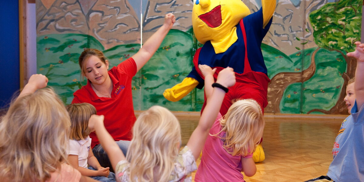 Eine Gruppe kleiner Kinder und ein Erwachsener sitzen auf dem Boden und heben die Arme, während eine Person in einem gelben Zeichentrickfilmkostüm in der Nähe in einem dekorierten Raum steht.