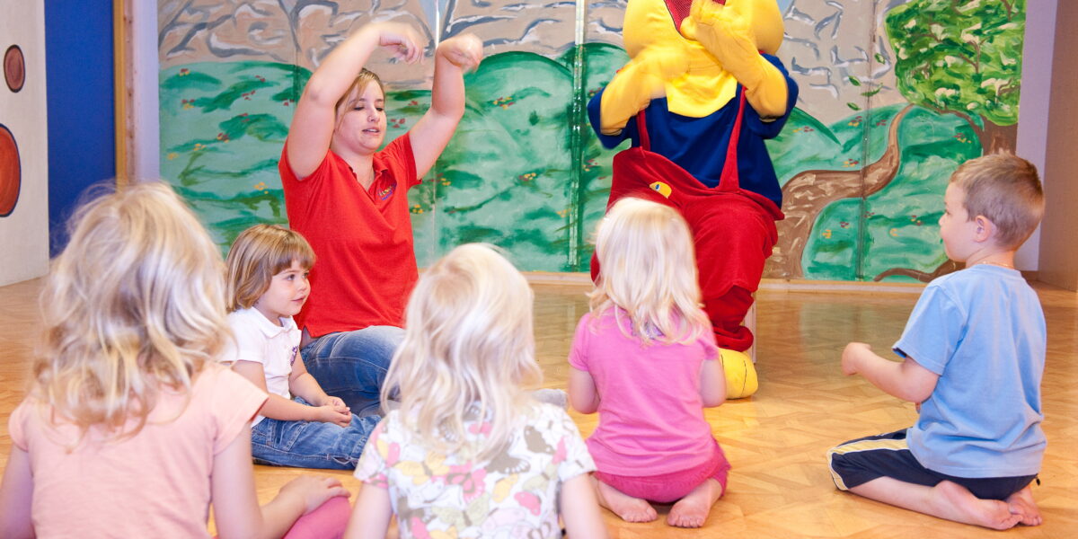 Eine Gruppe kleiner Kinder sitzt auf dem Boden und sieht sich einem Erwachsenen und einer Person in einem gelben Kostüm gegenüber, die in einem farbenfrohen Raum an einer interaktiven Aktivität teilnehmen.