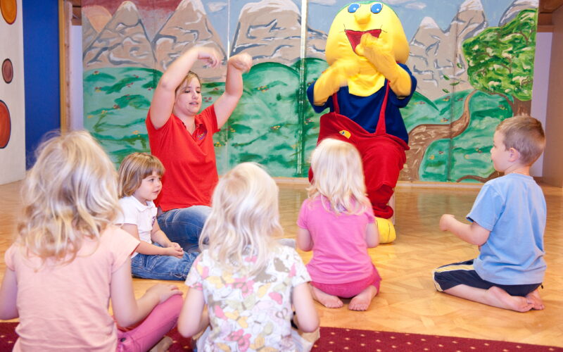 Eine Gruppe kleiner Kinder sitzt auf dem Boden und sieht sich einem Erwachsenen und einer Person in einem gelben Kostüm gegenüber, die in einem farbenfrohen Raum an einer interaktiven Aktivität teilnehmen.