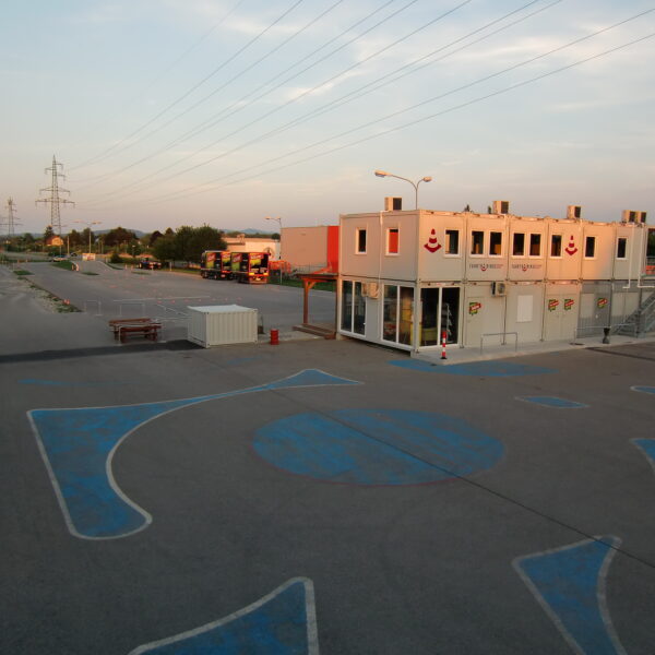 Leerer Parkplatz mit blauen Markierungen, ein kleines zweistöckiges Modulgebäude mit Beschilderung und ein paar Busse und Container im Hintergrund, unter Stromleitungen bei Sonnenuntergang.