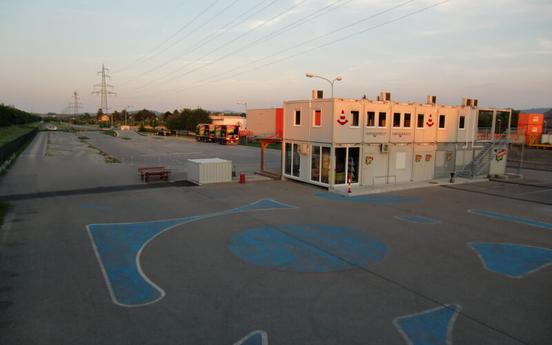 Leerer Parkplatz mit blauen Markierungen, ein kleines zweistöckiges Modulgebäude mit Beschilderung und ein paar Busse und Container im Hintergrund, unter Stromleitungen bei Sonnenuntergang.