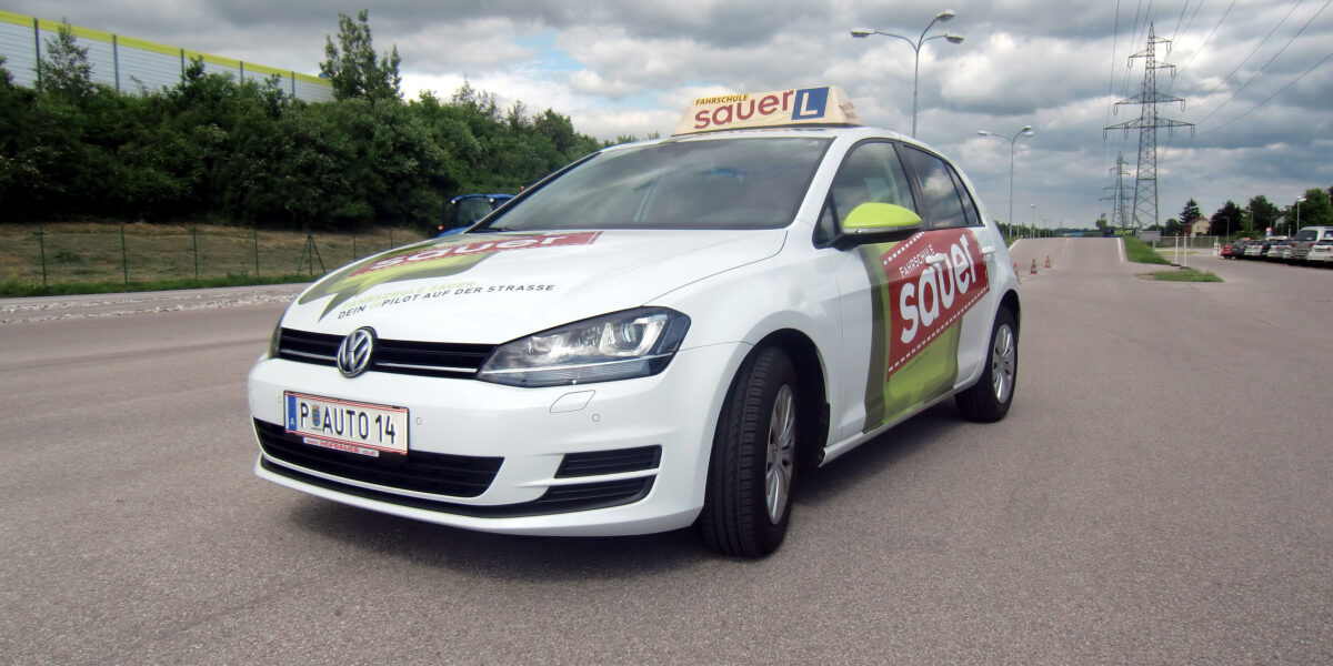 Ein weißer Volkswagen mit dem Schriftzug "sauer" und einem Schild für Fahrschüler parkt auf einer asphaltierten Straße unter einem bewölkten Himmel.