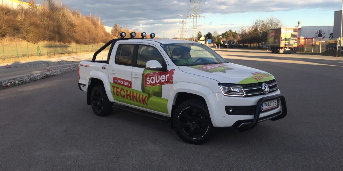 Ein weißer Volkswagen Amarok Pickup mit dem Schriftzug Sauer Technik" ist auf einer leeren asphaltierten Straße in der Nähe eines Industriegebiets geparkt.