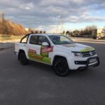 Ein weißer Volkswagen Amarok Pickup mit dem Schriftzug Sauer Technik" ist auf einer leeren asphaltierten Straße in der Nähe eines Industriegebiets geparkt.
