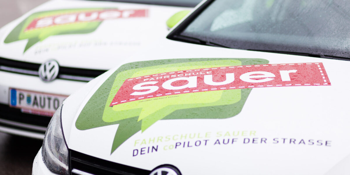 Zwei weiße Volkswagen mit den Logos "Fahrschule Sauer" und "Dein CoPilot auf der Straße" auf den Motorhauben, nebeneinander auf einer nassen Fläche geparkt.