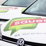 Zwei weiße Volkswagen mit den Logos "Fahrschule Sauer" und "Dein CoPilot auf der Straße" auf den Motorhauben, nebeneinander auf einer nassen Fläche geparkt.